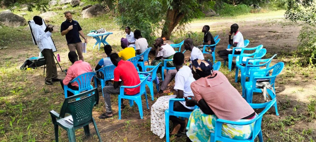 Udendørs klasseværelse. Deltagerne sidder i blå plastikstole og kigger mod de to undervisere
